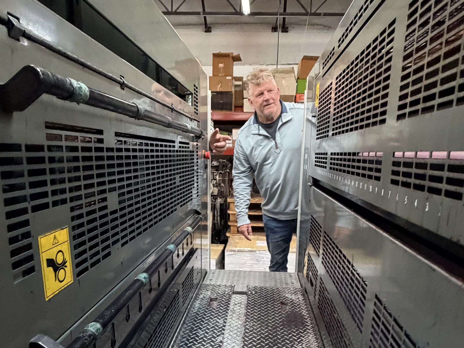 Randy Williams, the owner, leans out between two large printing presses on his shop floor, shelves of boxes behind him
